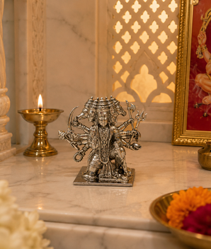 A silver metallic Lord Hanuman statue displayed on a marble altar alongside a burning brass oil lamp, a framed red deity painting, and incense sticks.