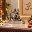 An ornate silver-toned Hindu deity statue on a marble altar, surrounded by a lit brass oil lamp, white flowers, and a bowl of marigolds, with a blurred Om symbol in the background.