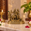An ornate gold-toned metal statue of Hindu deities and a cow on a decorative base, displayed on a marble surface with candles, flowers, and a potted plant in the background.
