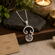 Tamil Om pendant brass silver finish on white ceramic plate with Tamil Om branded box, green plant and pen in background