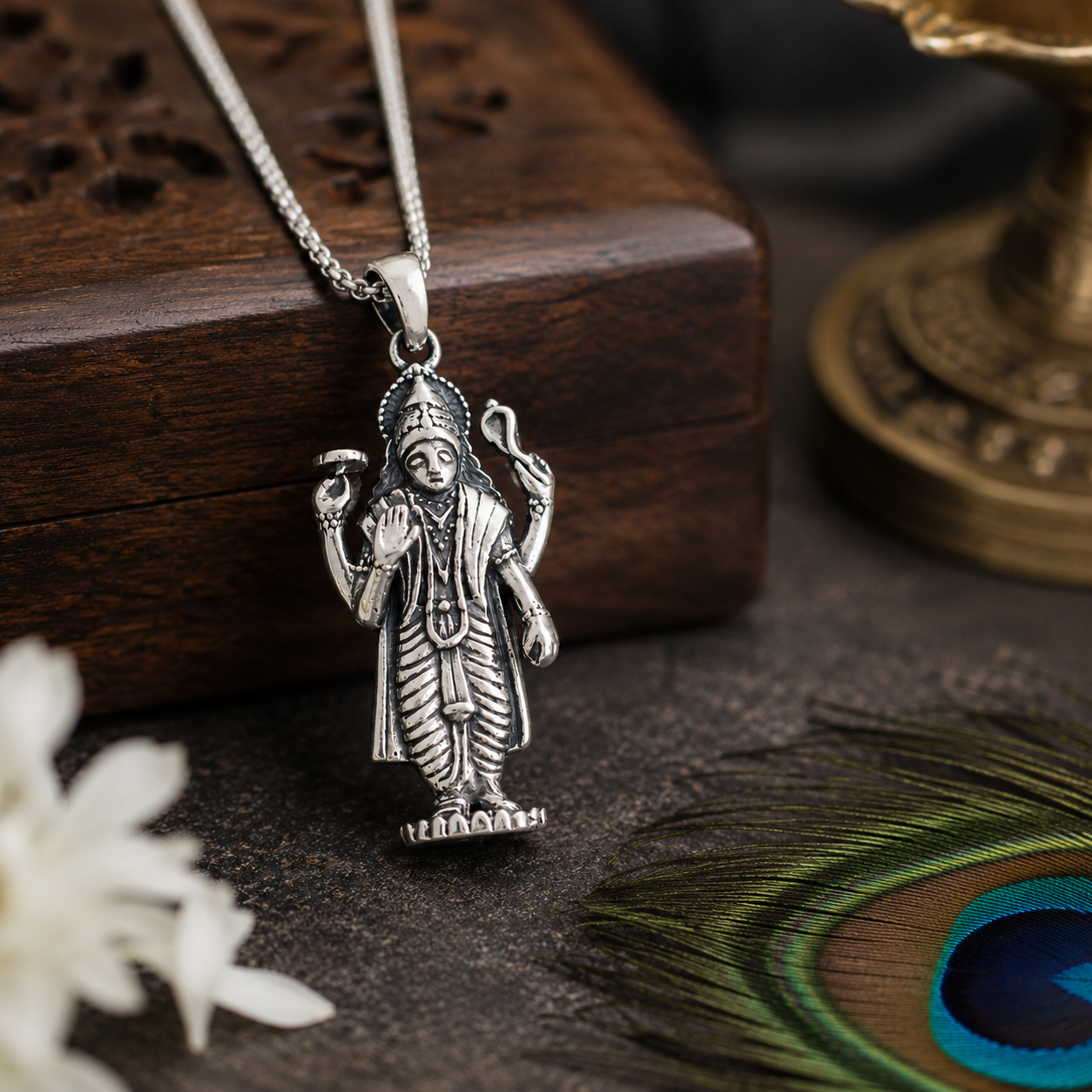 Lord Vishnu Ji pendant in brass silver finish resting against carved wooden block with peacock feather and brass lamp in background
