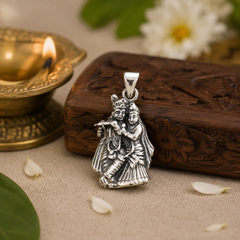 Radha Krishna pendant in brass antique silver finish resting on carved wooden box with lit brass diya, white jasmine flower and rose petals in warm devotional setting