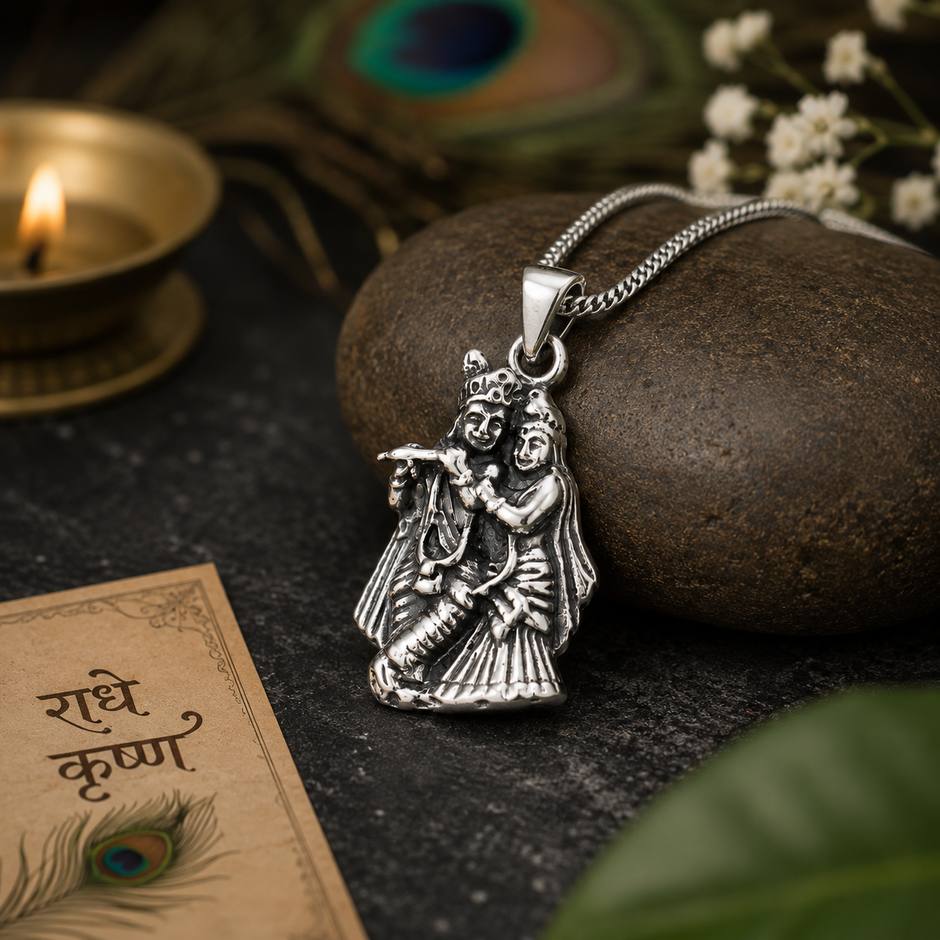 Radha Krishna pendant in brass antique silver finish on dark pebble stone with silver chain, Radhe Krishna Hindi calligraphy card, peacock feathers and lit diya in background