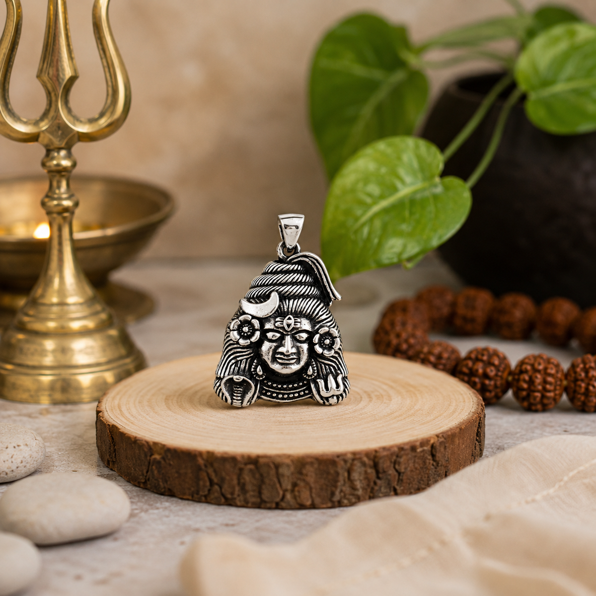 Mahadev Ji Shiv Ji face pendant in brass silver finish on wooden slice with brass trishul lamp, rudraksha bracelet, white pebbles and green leaves in background
