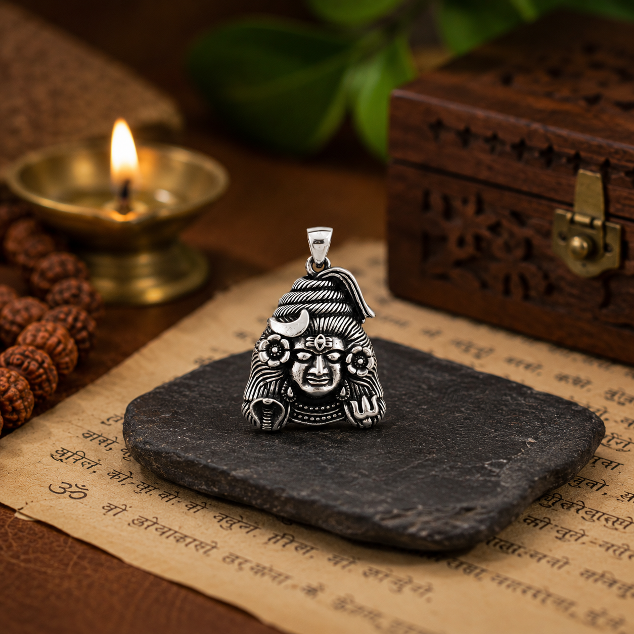 Mahadev Ji pendant in brass antique silver finish on dark slate stone on top of Sanskrit scripture with rudraksha mala, lit diya and carved wooden box in background