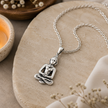 Gautam Buddha pendant in brass antique silver finish on silver chain resting on white marble stone with candle, white flowers and white linen in serene meditation setting