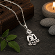 Gautam Buddha pendant in brass antique silver finish on silver chain on dark slate stone with carved wooden block, green tulsi leaves and lit amber candle in background