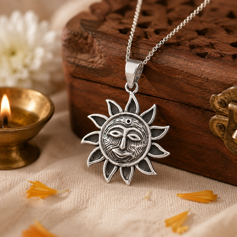 Surya Dev Sun God pendant in brass antique silver finish on silver chain resting on carved wooden jali box with lit brass diya and white chrysanthemum flowers and marigold petals