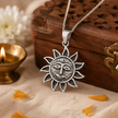 Surya Dev Sun God pendant in brass antique silver finish on silver chain resting on carved wooden jali box with lit brass diya and white chrysanthemum flowers and marigold petals