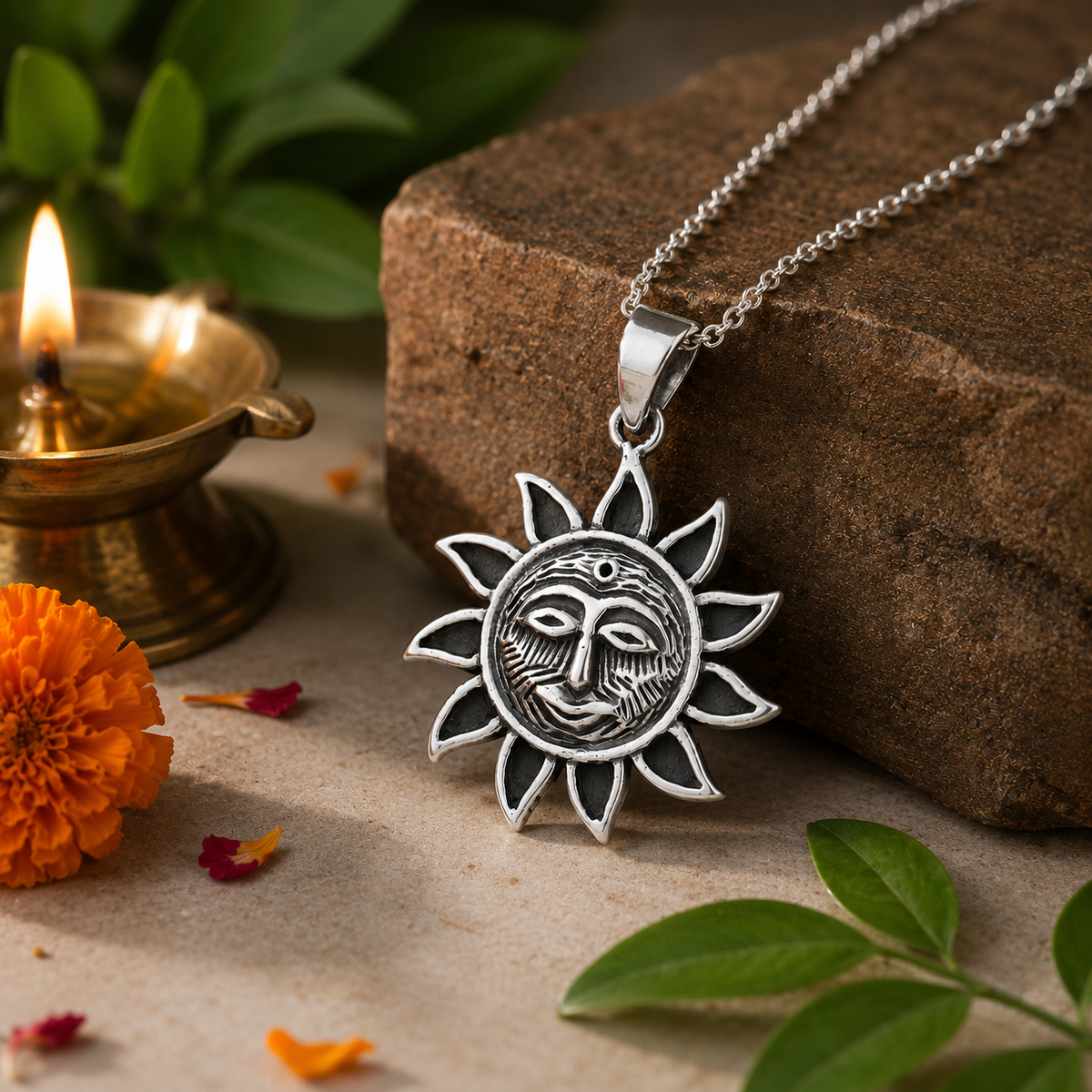 Surya Dev Sun God pendant in brass antique silver finish on silver chain leaning on brown sandstone rock with lit brass diya, orange marigold flower and green tulsi leaves