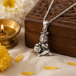 Laddu Gopal Baby Krishna pendant in brass antique silver finish on silver chain hanging from carved wooden jali box with lit brass diya and yellow chrysanthemum flowers and marigold petals on white linen