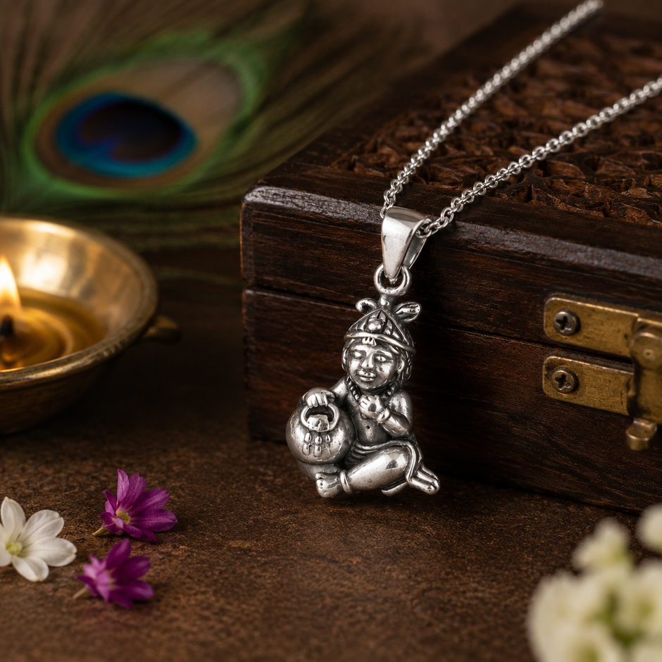 Laddu Gopal Baby Krishna pendant in brass antique silver finish on silver chain hanging from carved wooden treasure box with peacock feather, lit brass diya and purple and white wildflowers on dark brown background