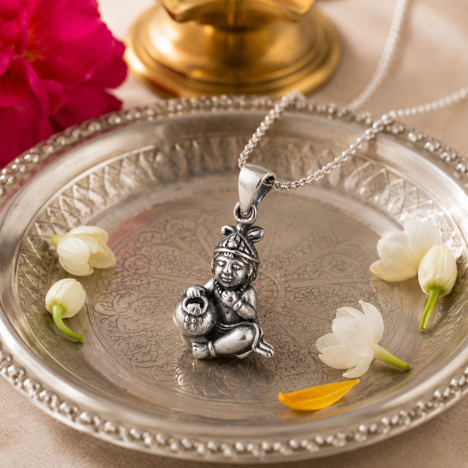 Laddu Gopal Baby Krishna pendant in brass antique silver finish on silver chain on engraved silver pooja thali plate with white jasmine flowers and marigold petals and brass lamp in soft warm devotional setting