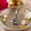 Laddu Gopal Baby Krishna pendant in brass antique silver finish on silver chain on engraved silver pooja thali plate with white jasmine flowers and marigold petals and brass lamp in soft warm devotional setting