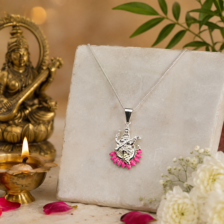 Maa Saraswati silver finish pendant with pink enamel lotus petals on silver chain leaning against white marble block with brass Saraswati idol, lit brass diya, pink rose petals and white baby breath flowers