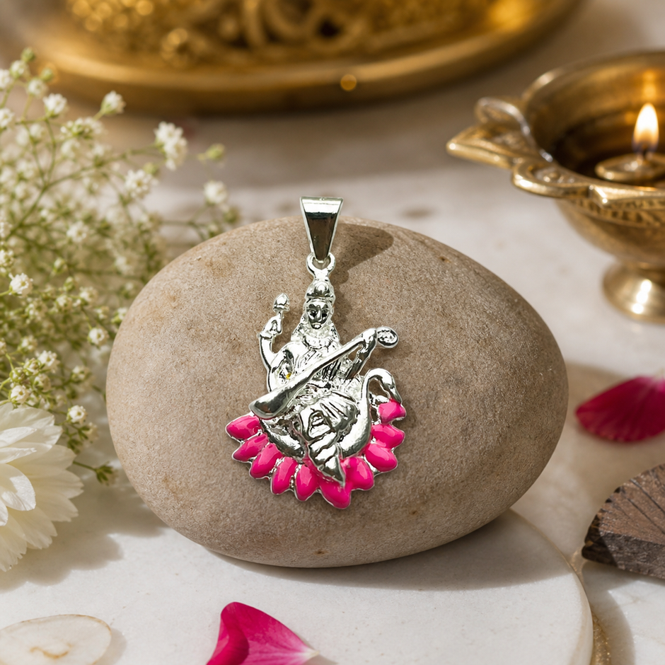 Maa Saraswati silver finish pendant with pink enamel lotus on silver chain hanging from beige velvet jewellery box with brass Saraswati idol, lit brass diya, white chrysanthemums, red rose and pink rose petals