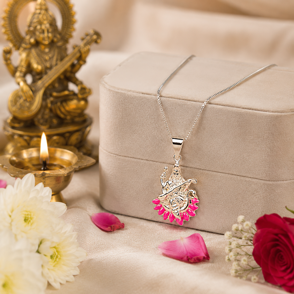 Maa Saraswati silver finish pendant with pink enamel lotus on silver chain hanging from beige velvet jewellery gift box on cream silk fabric with white chrysanthemums, red rose, pink petals and lit brass diya