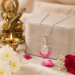 Maa Saraswati silver finish pendant with pink enamel lotus on silver chain hanging from beige velvet jewellery gift box on cream silk fabric with white chrysanthemums, red rose, pink petals and lit brass diya