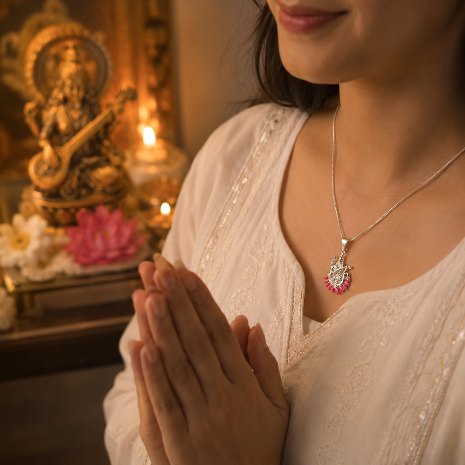 Woman in white embroidered saree wearing Maa Saraswati silver finish pink enamel lotus pendant on silver chain in Namaste prayer pose with brass Saraswati idol, lit diya and pink lotus in home temple background