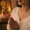 Woman in white embroidered saree wearing Maa Saraswati silver finish pink enamel lotus pendant on silver chain in Namaste prayer pose with brass Saraswati idol, lit diya and pink lotus in home temple background