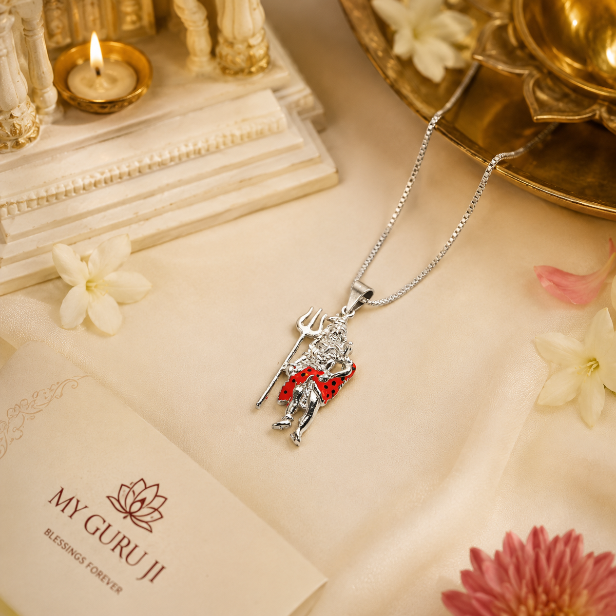 Mahadev Ji standing Shiva brass silver finish pendant with red enamel dotted dhoti on silver chain lying on white marble block with My Guru Ji branded card, white flowers, pink chrysanthemum and lit tealight candle on cream linen background