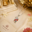 Mahadev Ji standing Shiva brass silver finish pendant with red enamel dotted dhoti on silver chain lying on white marble block with My Guru Ji branded card, white flowers, pink chrysanthemum and lit tealight candle on cream linen background