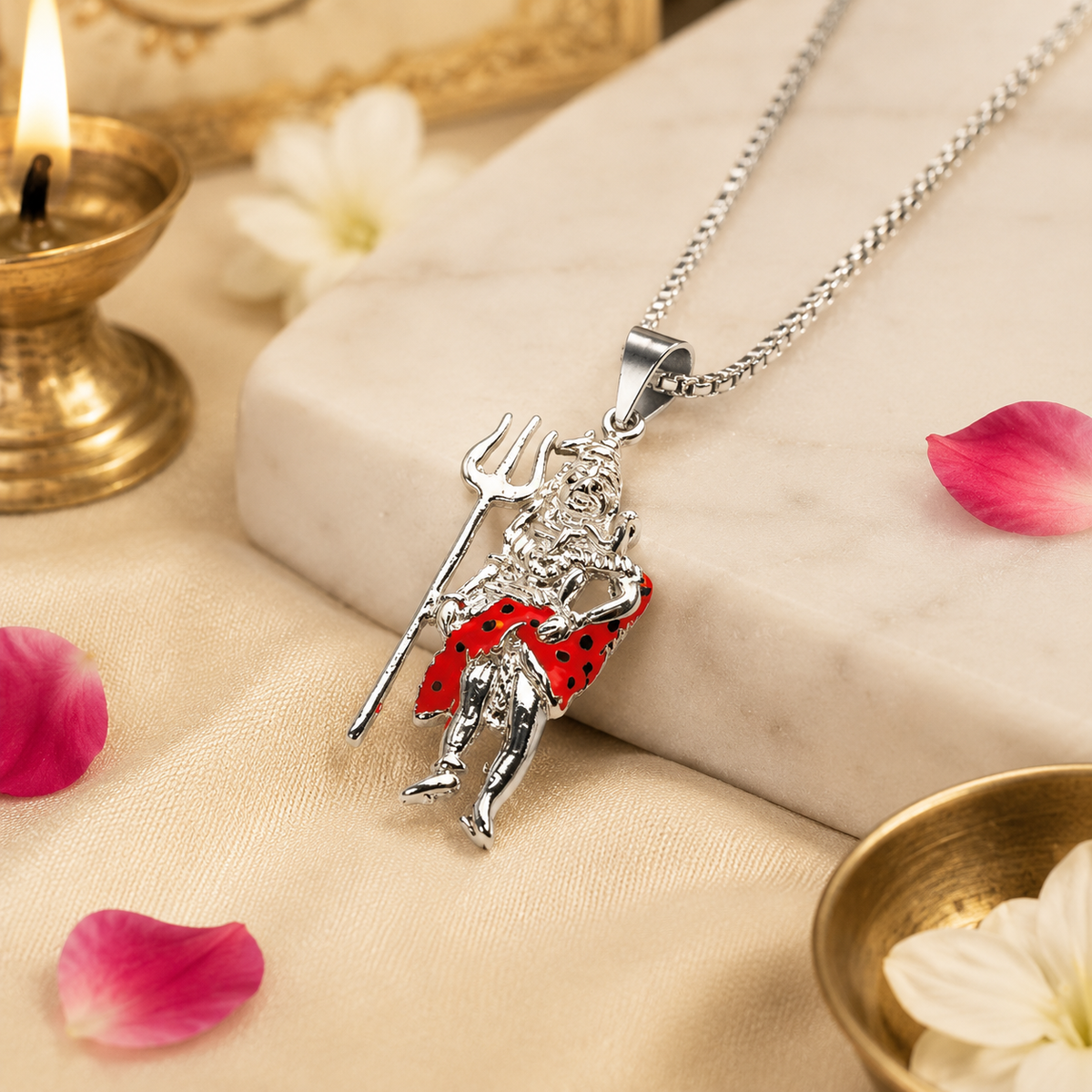 Mahadev Ji standing Shiva brass silver finish pendant with red enamel dotted dhoti on silver chain lying on white marble slab with lit brass diya, pink rose petals and white jasmine flowers on cream satin background close-up detail shot