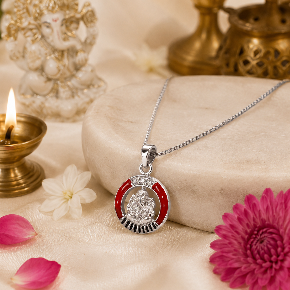 Ganesh Ji brass silver finish pendant with red enamel ring CZ crown stones and black sunburst rays on silver chain on white marble slab with lit brass diya, white Ganesha idol, pink chrysanthemum flower and rose petals on cream background