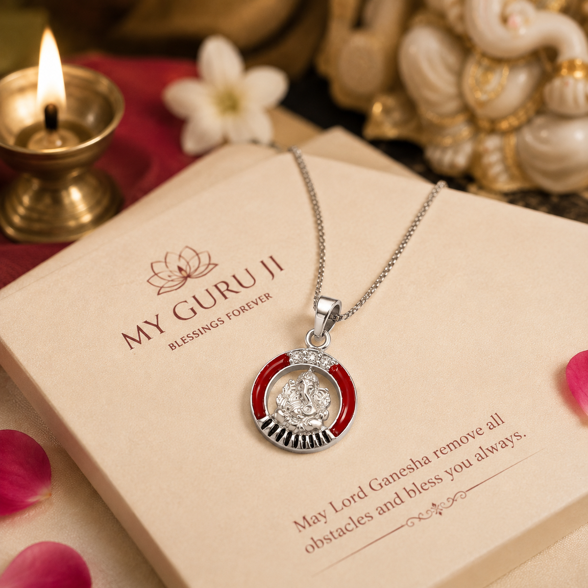 Ganesh Ji brass silver finish pendant with red enamel ring CZ crown and black sunburst rays on silver chain on My Guru Ji branded cream card reading may Lord Ganesha remove all obstacles and bless you always, with white Ganesha idol, lit diya, white jasmine flower and rose petals