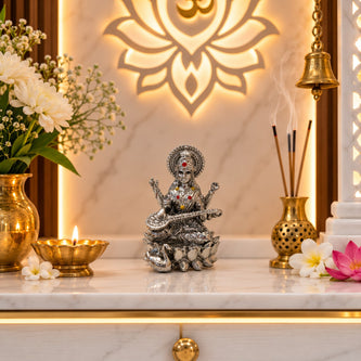My Guru Ji silver finish brass Maa Saraswati idol seated on a lotus with a veena, displayed in a home temple setting.